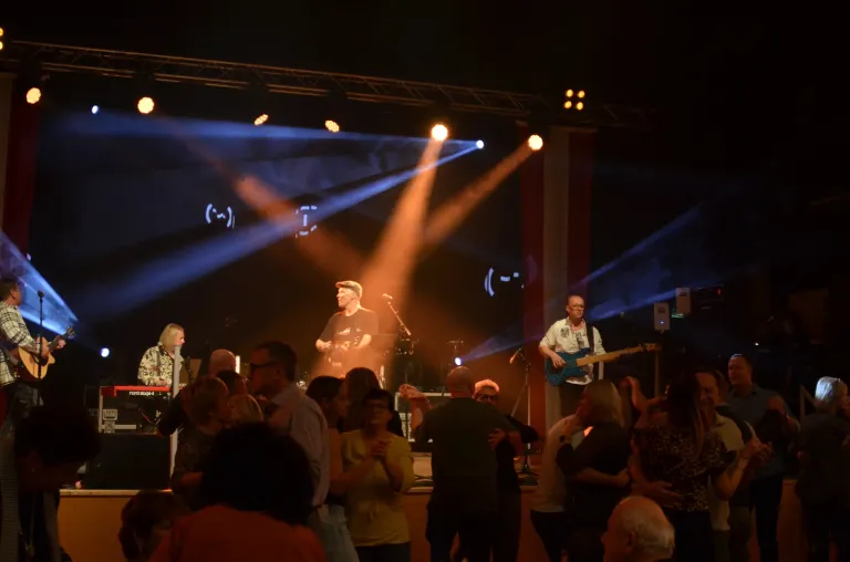 A band performs on stage under blue and yellow lights. The lead singer plays the guitar, and the audience dances and claps.