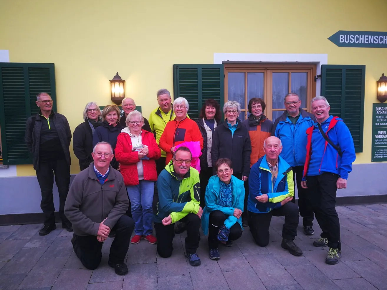 A group of adults, some wearing glasses, are smiling and posing for a photo outside a building with yellow walls and green shutters.