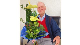 An elderly man sits in a chair holding a bouquet of green flowers. He wears a red sweater and a blue cardigan.
