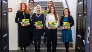 Six women stand on a staircase, holding colorful books. They wear winter clothing and seem to be posing for a photograph.