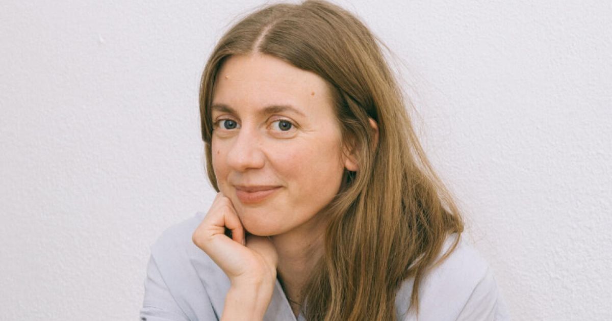 A woman with long brown hair and a light blue shirt is smiling at the camera, with her hand on her cheek.