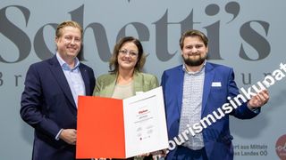 Three individuals pose with a diploma; one woman holds it, flanked by two men, all smiling in front of a wall with a large letter 'i'.