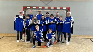A group of people, wearing blue jerseys, are posing for a photo in a sports hall with a wooden floor. They are holding hockey sticks and standing in front of a red and white goalpost.