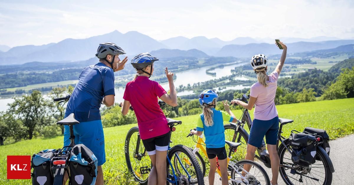 A family of four, two adults and two children, stands on a grassy hill with their bicycles, taking in the scenic view of mountains and a river in the distance.