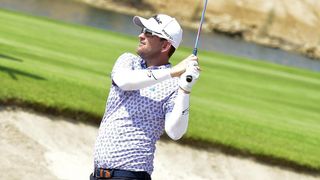 A golfer in a white hat and sunglasses swings his club on a golf course with a green field and a water body in the background.