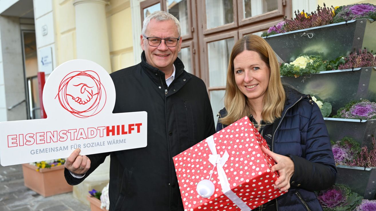 A man and a woman stand smiling in front of a building, holding a sign and a wrapped gift. The sign reads 'Stadthilfe' and the gift is red with white polka dots.