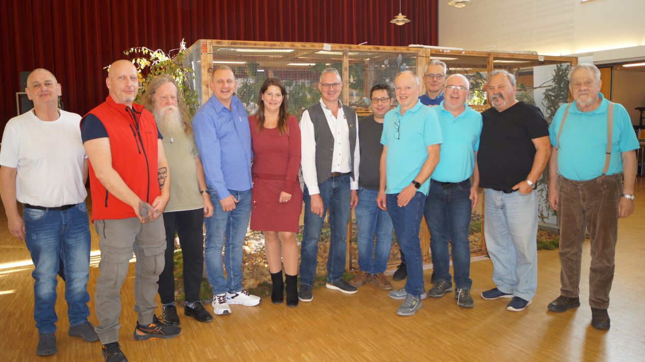 A group of people are posing for a photo in a room with wooden flooring. Some are wearing glasses, and others are smiling. There are bird cages behind them.