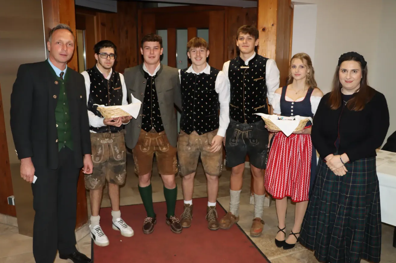 A group of young people dressed in traditional Bavarian clothing stand together in a room. They are smiling and posing for a photo.