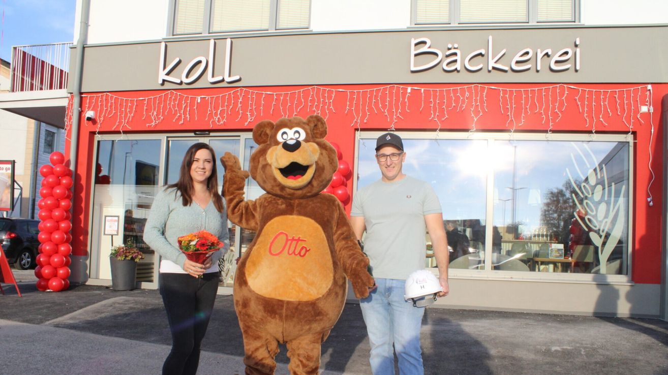 A woman and a man stand outside a bakery shop with a mascot. The mascot is a bear wearing a red Otto costume. The man is wearing a helmet and holding a bouquet. Behind them are red balloons.
