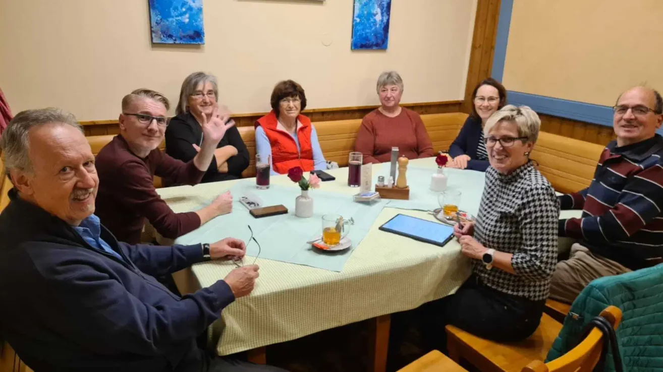 A group of people sit at a table in a restaurant. There are flowers, drinks, and a tablet on the table. Everyone is smiling.