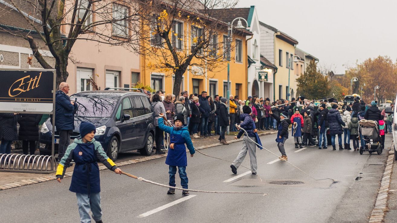 Kinder ziehen eine Seil in der Straße, während eine Menge zuschaut. Gebäude in verschiedenen Farben säumen die Straße.