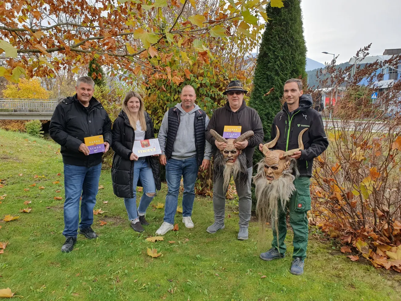 Five people stand in a garden with autumn leaves, smiling for the camera. One man wears a costume with horns. Each holds a book, and one has a purple cover.