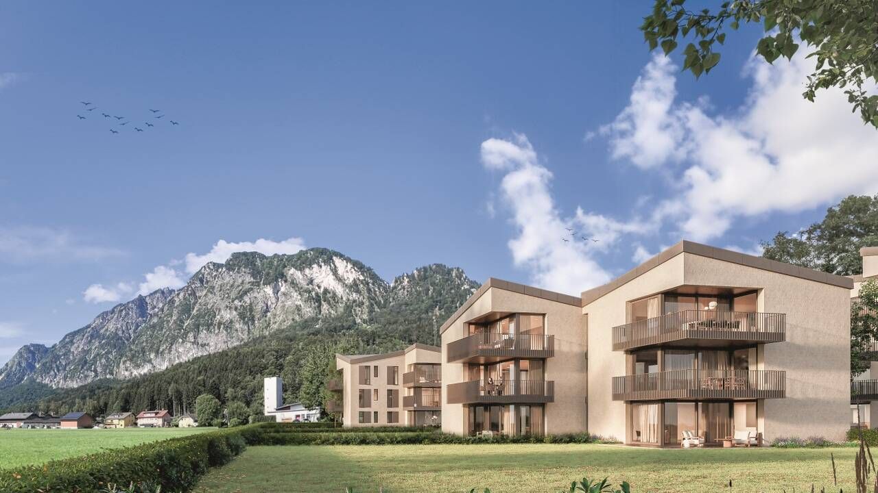 A modern apartment complex is situated in a green valley with a backdrop of mountains under a blue sky with clouds.