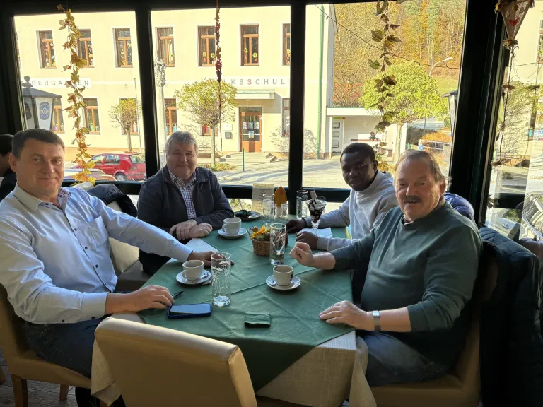 Vier Männer sitzen um einen Tisch in einem Restaurant und lächeln. Jeder hat eine Tasse vor sich. Hinter ihnen zeigt ein großes Fenster einen Blick auf Bäume und Gebäude.