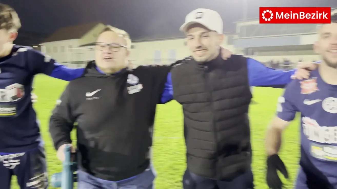 Zwei Männer stehen auf einem Fußballfeld. Einer trägt eine schwarze Jacke, der andere eine schwarze Weste. Beide lächeln.
