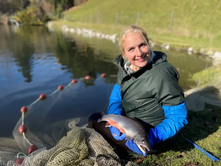 Eine Frau in einer grünen und blauen Jacke, die einen Fisch hält und lächelt, am See.