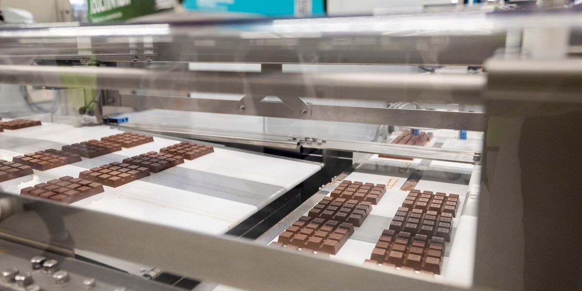 Eine industrielle Schokoladenproduktionsmaschine verarbeitet Schokoladentafeln auf einem Förderband. Die Maschine verfügt über Edelstahlschalen mit Schokoladen und einer transparenten Abdeckung.