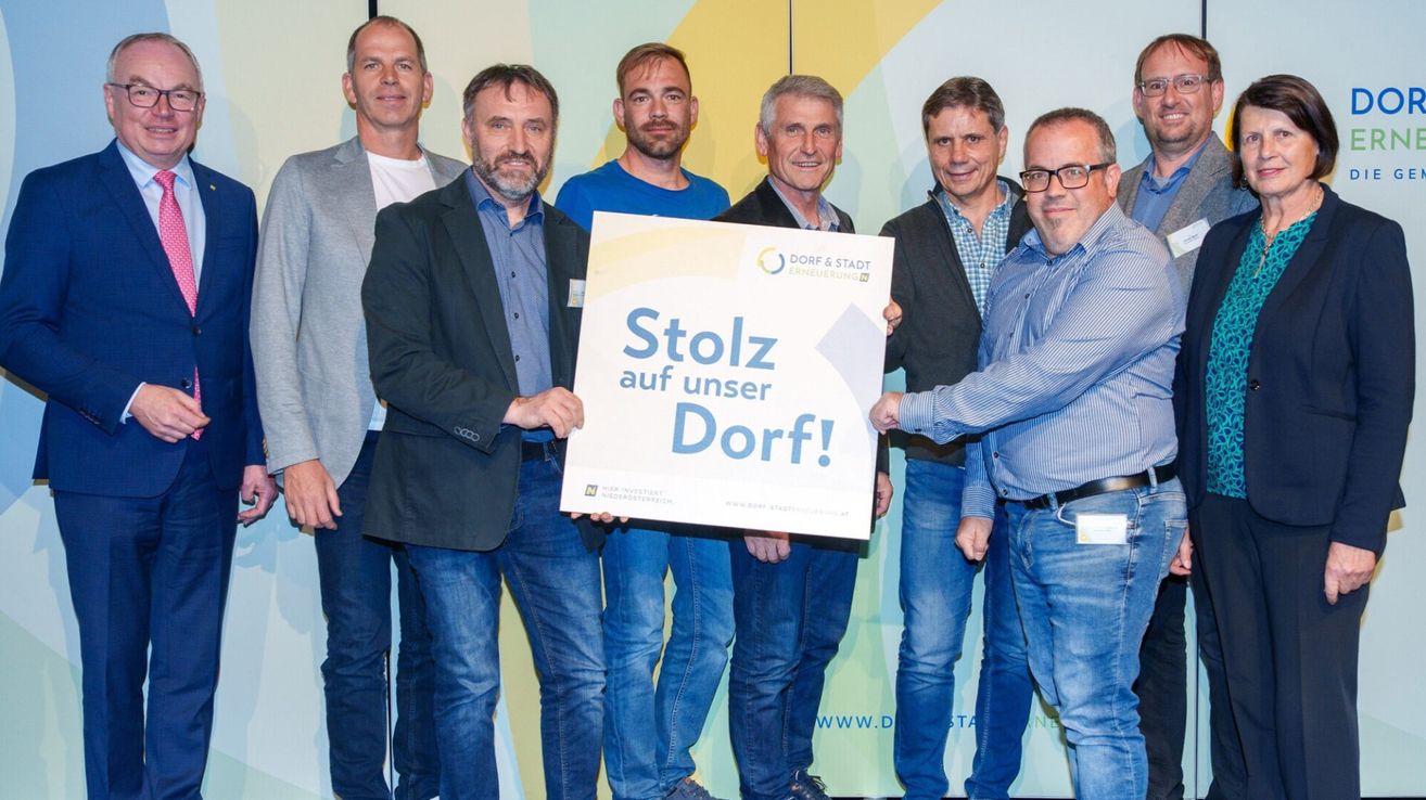 A group of men standing together holding a sign that says Stolz auf unser Dorf! They are smiling and seem to be at an event.