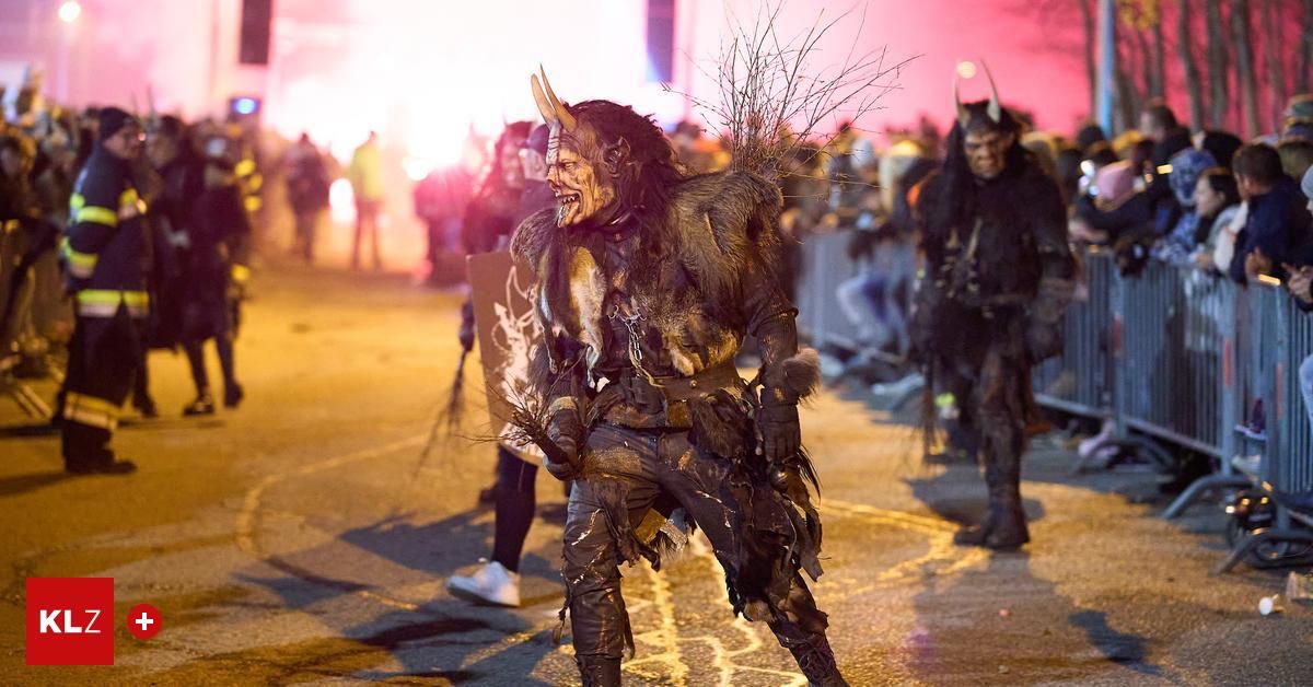 A person in a horned costume walks down a street at night with others, holding a shield, while a crowd watches.