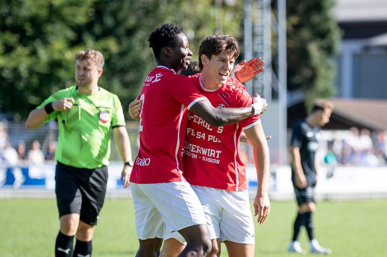 Two soccer players in red jerseys hug each other while the referee stands nearby on the field.