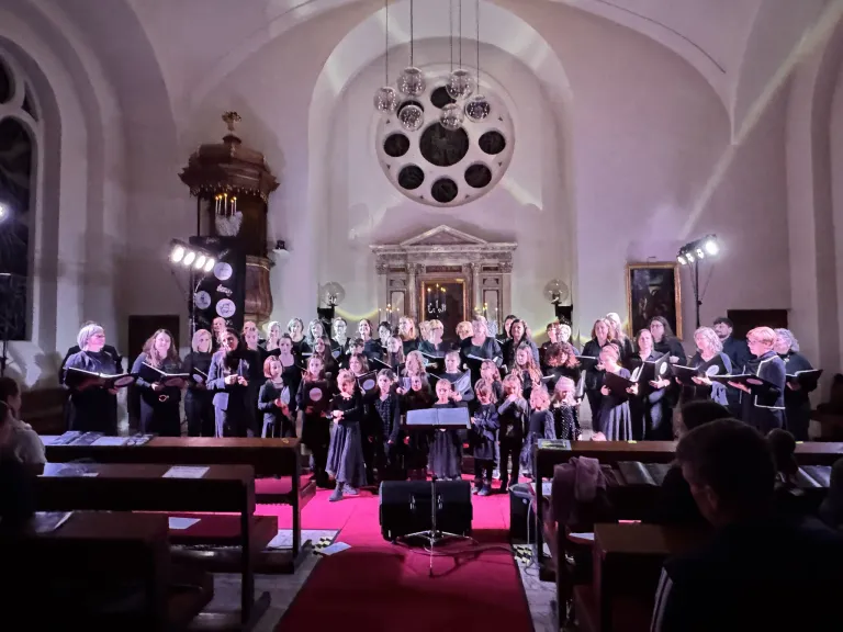 Ein Chor in schwarz gekleidet singt in einer Kirche und hält Bücher. Das Publikum sitzt auf Bänken. Ein Lautsprecher steht auf der Bühne mit einem roten Teppich.