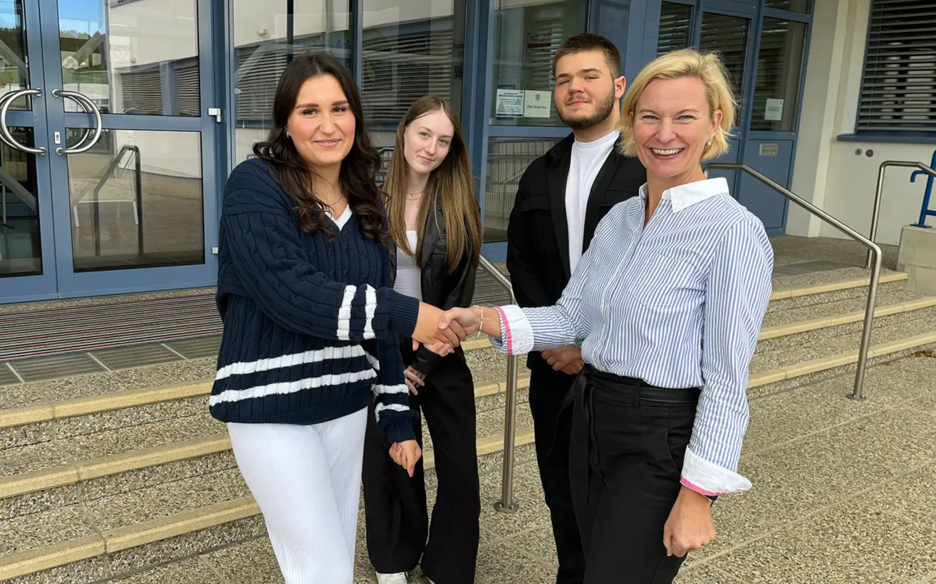 Four individuals stand on a staircase, smiling. A woman in a blue sweater shakes hands with another woman in a striped shirt. Behind them, two others stand, one with a beard. The background features a glass wall and a building.