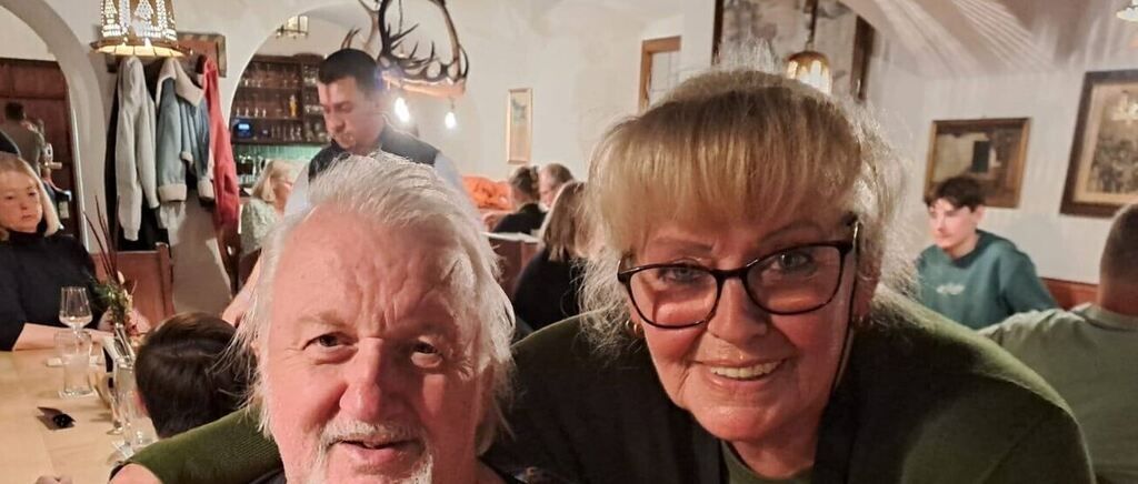 An elderly man with a white beard and mustache poses with a smiling older woman wearing glasses inside a crowded room. Behind them, several people sit at tables. A man stands near a shelf with bottles and a deer head mounted on the wall.