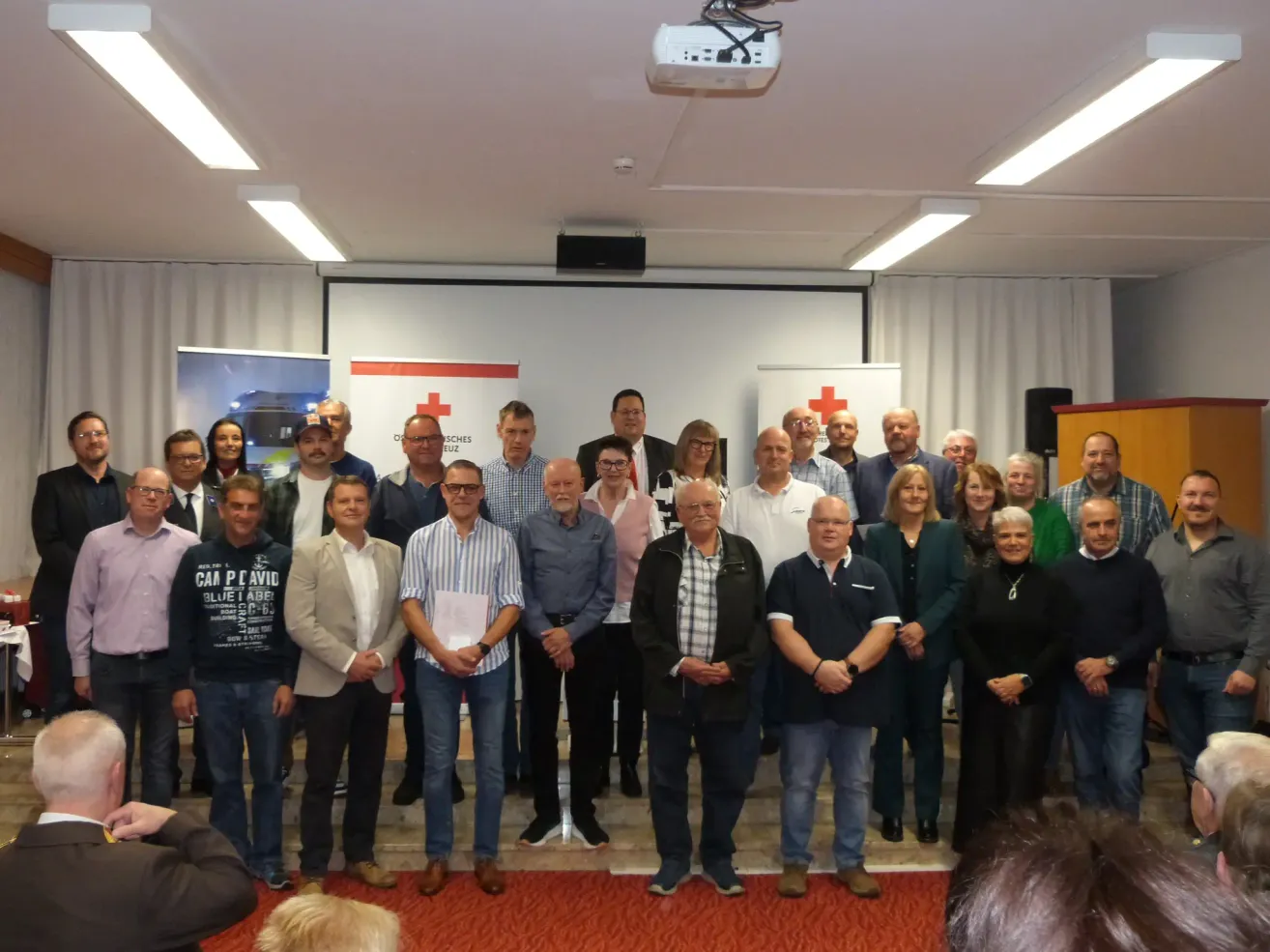 A group of people stand in front of a red cross banner in a room, some smiling and others holding papers. A projector and speakers are mounted on the ceiling.