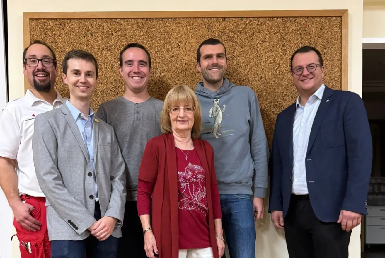Five people stand together in front of a wall, smiling. A woman with glasses and a necklace is in the center. On the left, a man in a gray jacket and a man in a gray shirt. On the right, a man in a blue suit and a man in glasses.