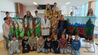 A group photo of students and teachers posing in a classroom, with two large art pieces in the background.