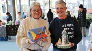 Two women stand together in a store, one holding a colorful bag and the other holding a small holiday decoration.