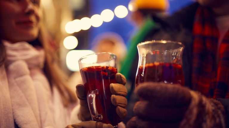 Two hands hold glasses of red liquid with blurry people and lights in the background.