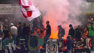 A crowd of fans at a sports event, some standing on barriers and holding flags, with smoke in the background. A man is holding a torch, and there is a banner with the number 25.
