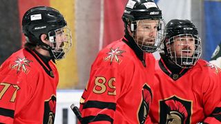 Three hockey players in red jerseys and helmets stand on an ice rink. One player wears the number 89.