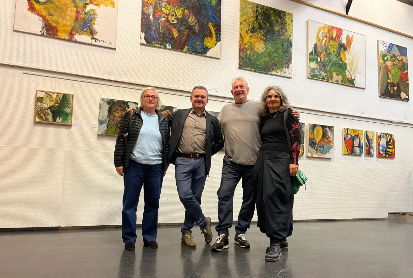 Four people are standing together in an art gallery, smiling, with a variety of paintings displayed on the walls behind them.