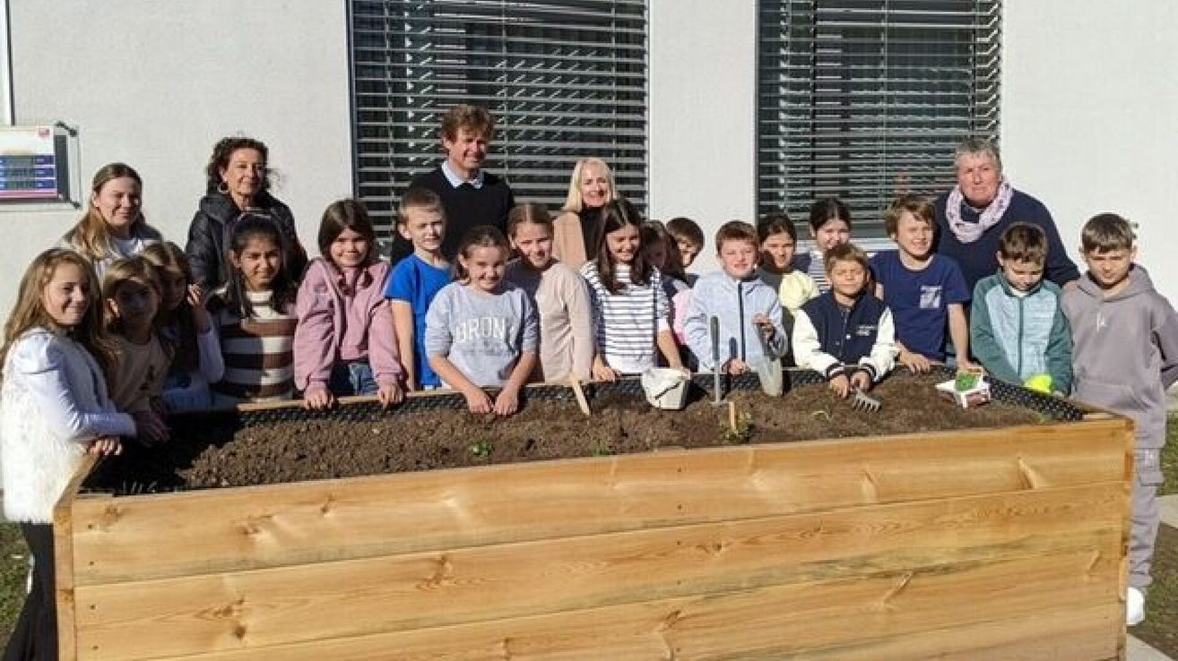 A group of children and adults are gardening in a wooden planter. They hold gardening tools and stand in front of a building with blinds.