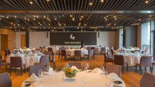 A large banquet hall with multiple tables and chairs, adorned with white tablecloths and flowers. A large sign reading 'The Resort' is mounted on the wall.