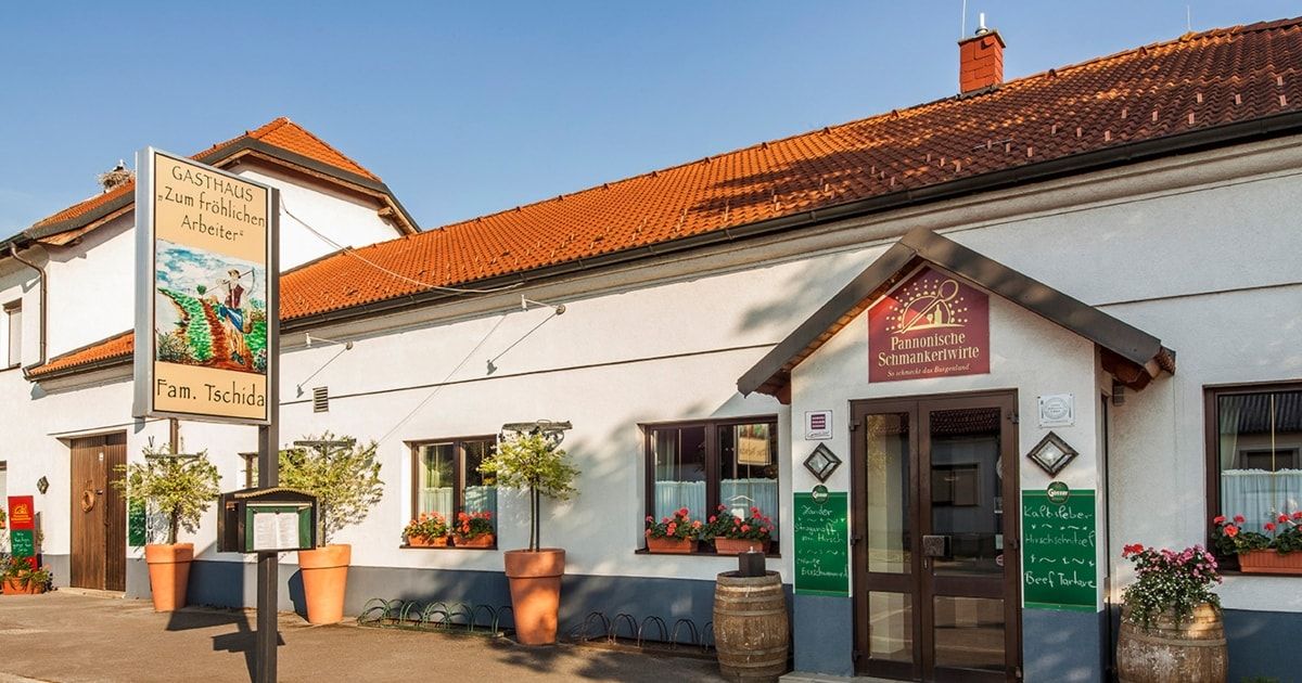 The exterior of a traditional German building with a red roof, a sign reading Pannonische Schmankerlwirte, a wooden barrel near the entrance, and plants in pots.