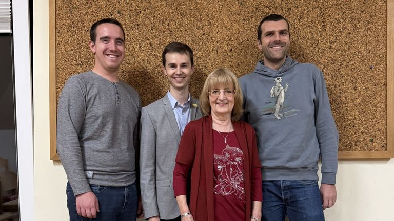 Four individuals stand close together in front of a textured brown wall, smiling for a photo. A woman in a red top and cardigan, and three men in gray and blue clothing.