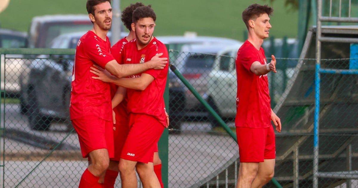 Four soccer players in red uniforms celebrate on a field. Behind them, a fence and parked cars are visible.