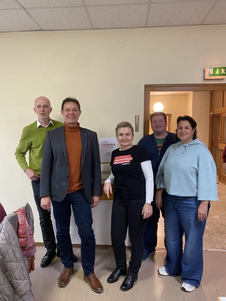 Five individuals are standing together in a room, smiling for a picture. A woman in the middle wears a shirt that says 'Ich Konnen Kindermut Schaffini'. Behind them is a wall with a sign and an emergency exit sign.
