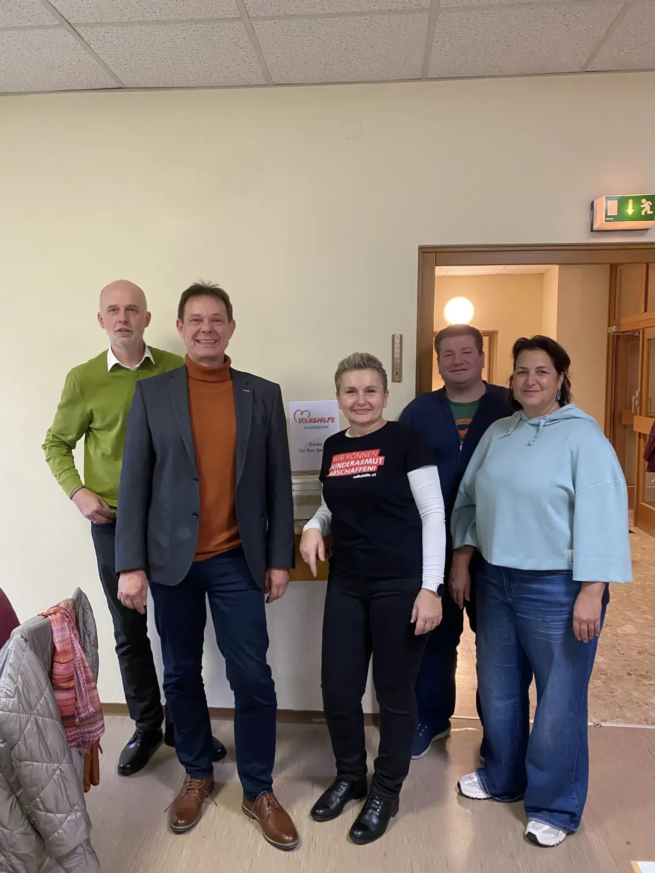 Five individuals are standing together in a room, smiling for a picture. A woman in the middle wears a shirt that says 'Ich Konnen Kindermut Schaffini'. Behind them is a wall with a sign and an emergency exit sign.