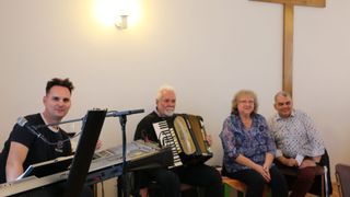 An older man plays an accordion in a room with a white wall and a woman sitting next to him smiling.