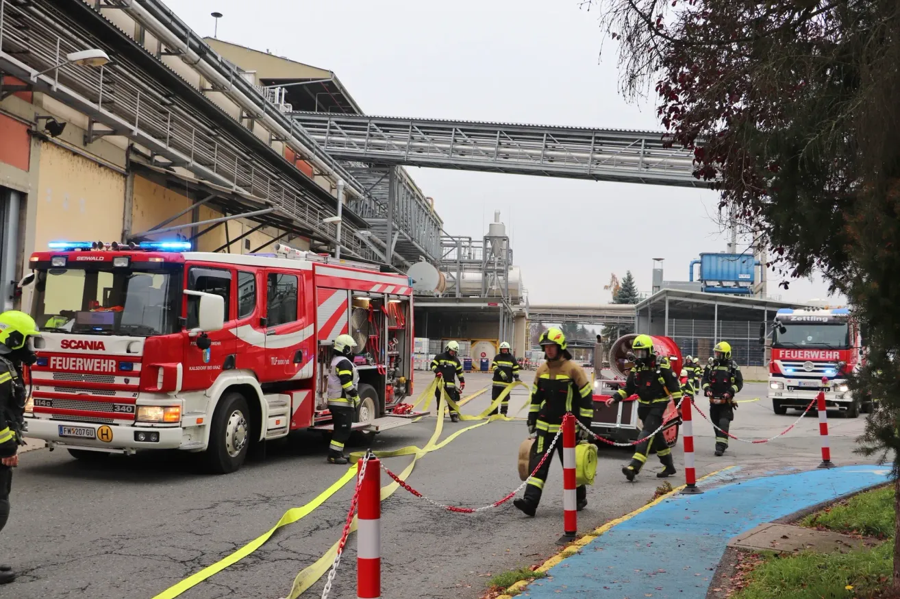 Firefighters in protective gear are stationed in front of a fire truck at an industrial site, connected by yellow ropes. The scene includes buildings and trees.