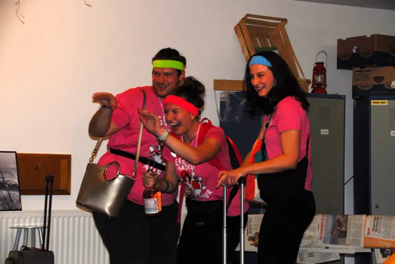Drei Personen in rosa T-Shirts und Stirnbändern lachen gemeinsam. Sie haben eine Flasche und eine Handtasche. Ein weißer Heizkörper und ein Holzkasten sind im Hintergrund.