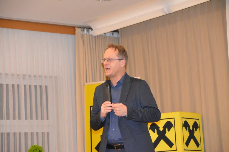 A man is giving a speech into a microphone in front of a yellow sign with an X symbol. He wears glasses and a suit. A curtain and white blinds are in the background.
