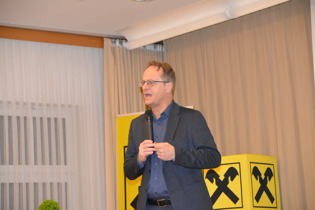A man is giving a speech into a microphone in front of a yellow sign with an X symbol. He wears glasses and a suit. A curtain and white blinds are in the background.