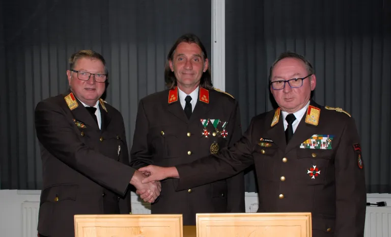 Drei Männer in Militäruniformen schütteln sich die Hände. Der Mann in der Mitte trägt mehrere Medaillen. Der Mann auf der linken Seite trägt eine Brille. Der Mann auf der rechten Seite trägt eine Brille und eine Krawatte.