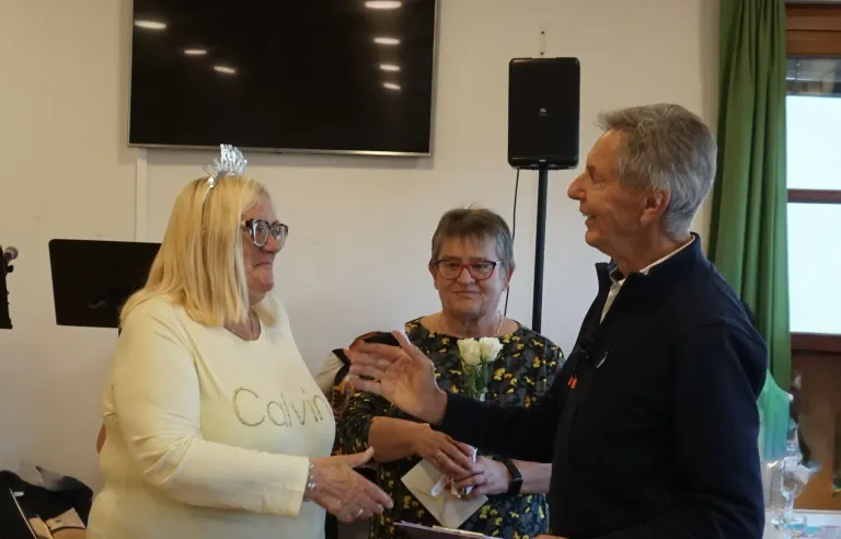 Three people stand in a room, two women and a man. The woman on the left wears a white shirt with the word Calvin. The woman in the middle holds a bouquet of flowers. The man smiles.