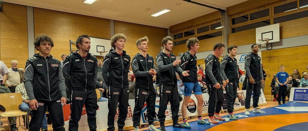 Eight male athletes stand in a line in a gym, all wearing matching black uniforms. They seem to be preparing for a competition. Behind them, there are people sitting on the bleachers. The wall has a basketball hoop and a banner.